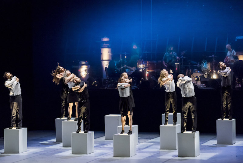 Dancers stand on pedestals with their arms crossed over their chests. The band is in the background.